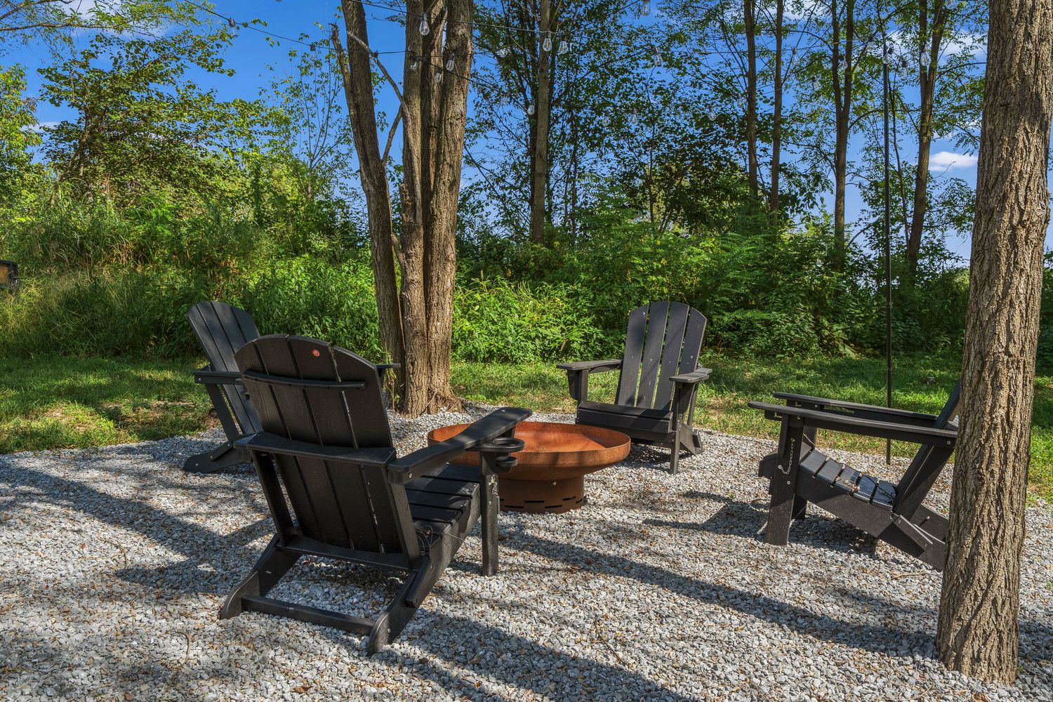 Adirondack chairs around a fire pit on a gravel pad under the trees.